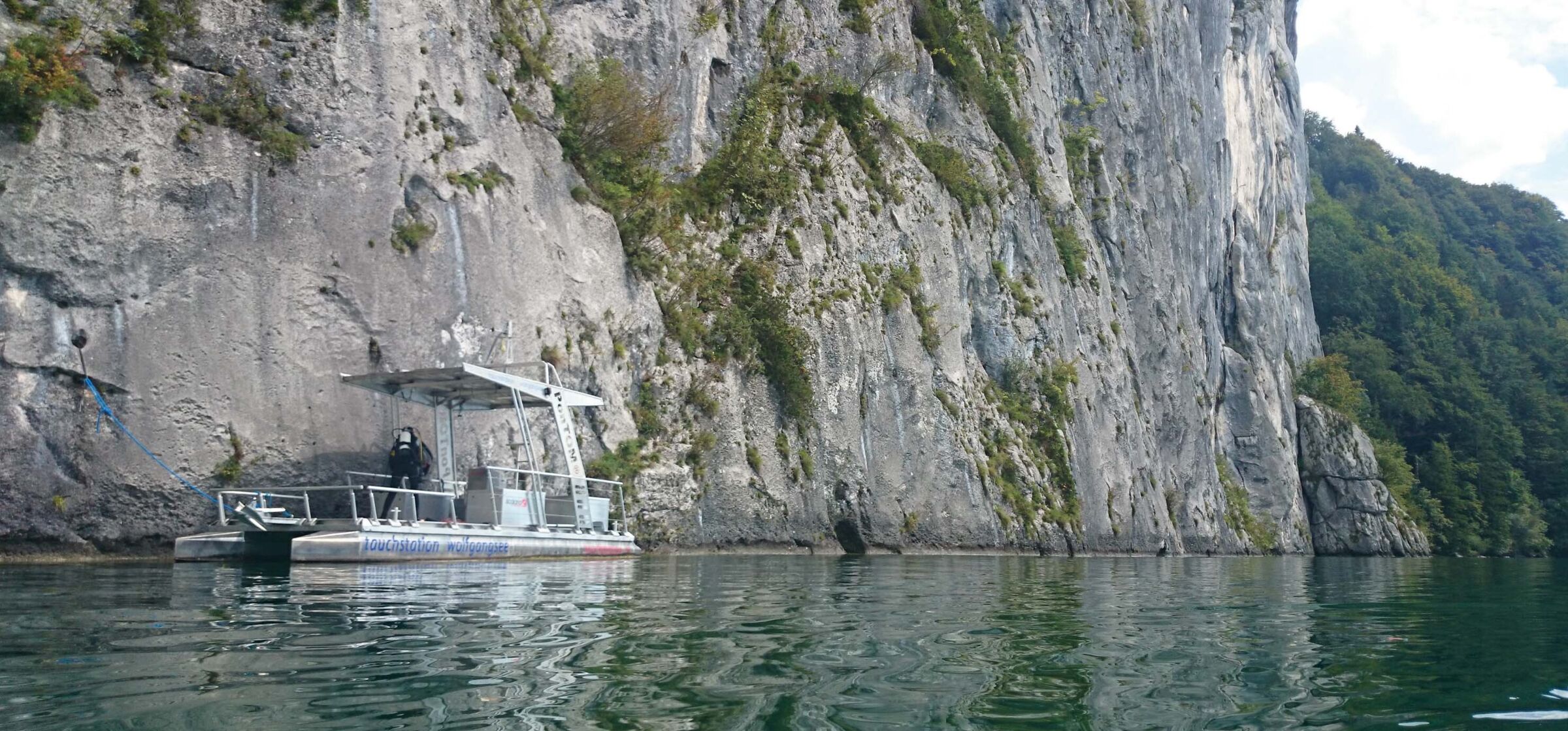 Taucher auf einem Boot im Wolfgangsee vor einer riesigen Felswand bereit zum Los tauchen