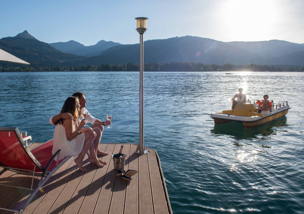 Verliebtes Pärchen mit einer Flasche Sekt auf einer Sonnenliege direkt am Wolfgangsee und zwei Tretboot fahrende Personen im Hintergrund