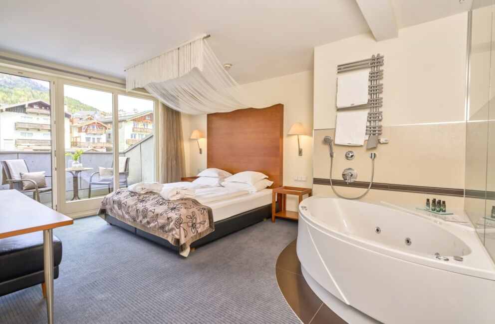 Zimmer im Romantik Hotel Im Weissen Rössl am Wolfgangsee mit Doppelbett, Whirlpool Badewanne, offenem Badbereich, Schreibtisch, Sofa und Balkonzugang.
