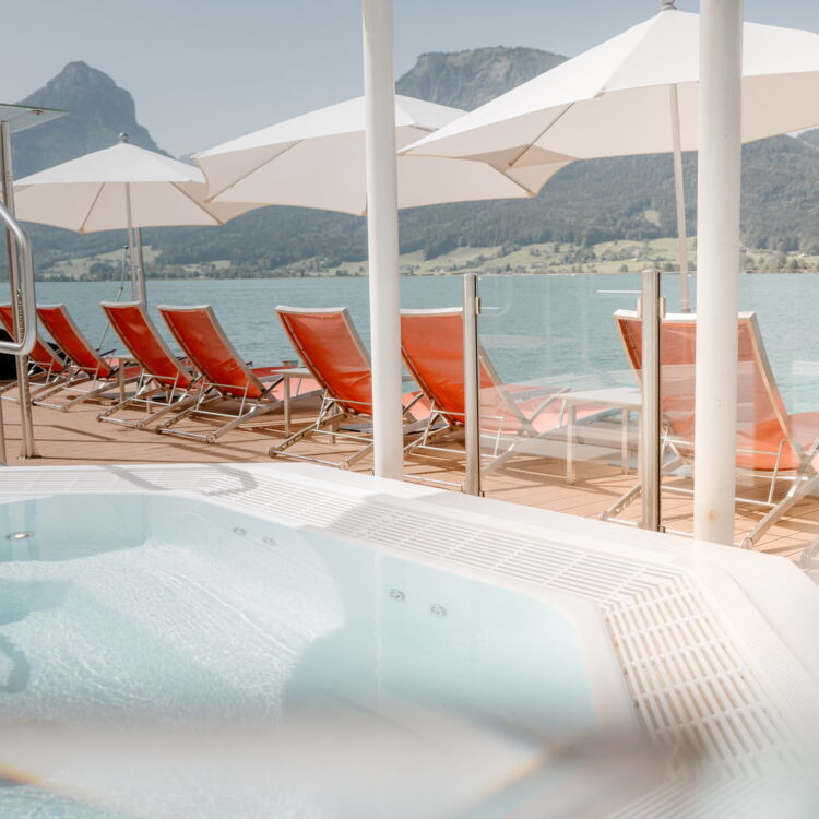 Beim Wellness am Wolfgangsee entspannen Sie am schwimmenden Whirlpool. Auf dem Holzdeck befinden sich rote Liegestühle und Sonnenschirme. Im Hintergrund sieht man den Wolfgangsee und die wunderschöne Bergkulisse.