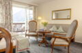 Sitzbereich im Romantik Hotel Im Weissen Rössl am Wolfgangsee mit Sofa, zwei Sesseln, rundem Tisch, Spiegel, floralen Vorhängen und Balkonzugang mit Blick auf den Wolfgangsee und die Berglandschaft.
