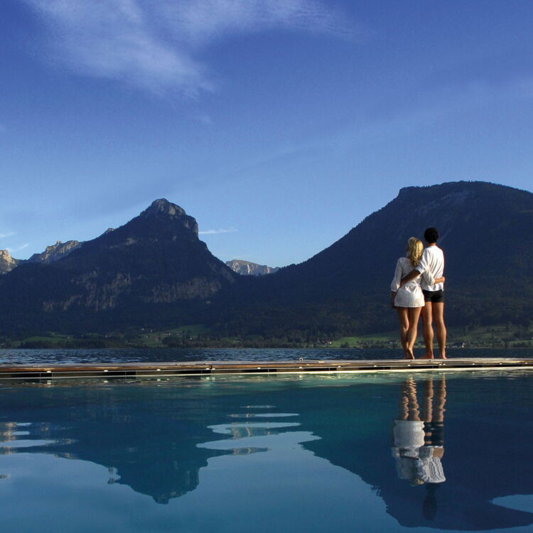 A couple stands arm in arm on a footbridge and looks out over the Wolfgangsee lake