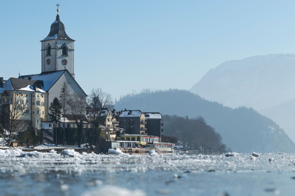 Blick vom Wolfgangsee aus auf das Romantik Hotel Im Weissen Rössl mit Eis und Schnee auf dem Wasser. Im Hintergrund ist die Kirche und bewaldete Hügel zu sehen.