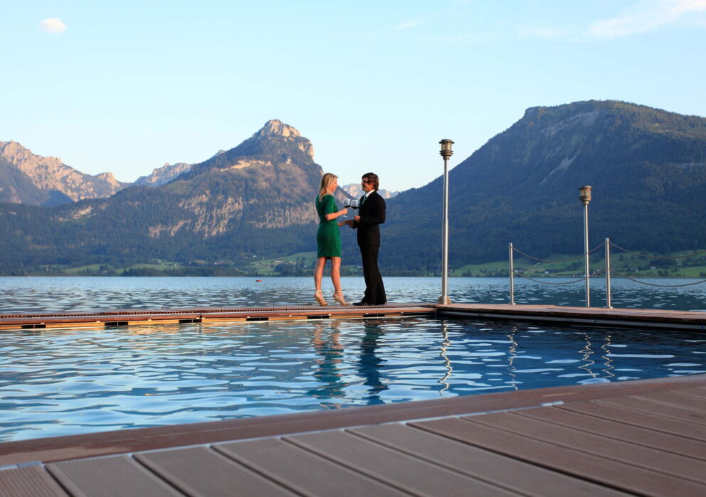 Schick gekleidetes Pärchen mit Wein anstoßend auf dem beheizten Seebad am Wolfgangsee mit spektakulärer Kulisse