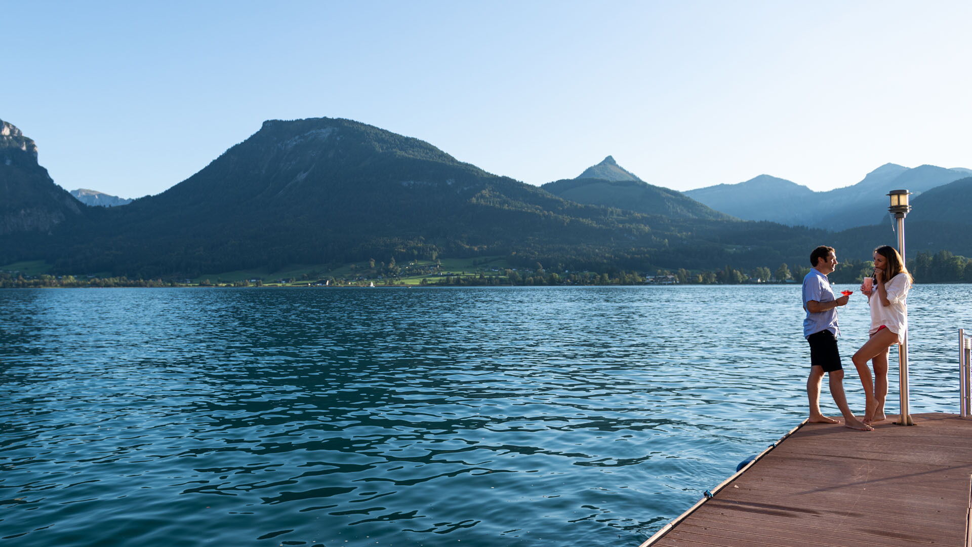 Ein Pärchen steht auf einem Steg am Wolfgangsee und trinkt Cocktails