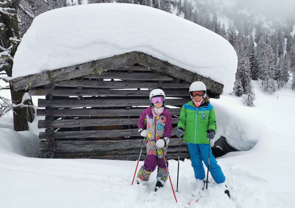 Zwei Mädchen mit Ski vor einer tief verschneiten Futterhütte