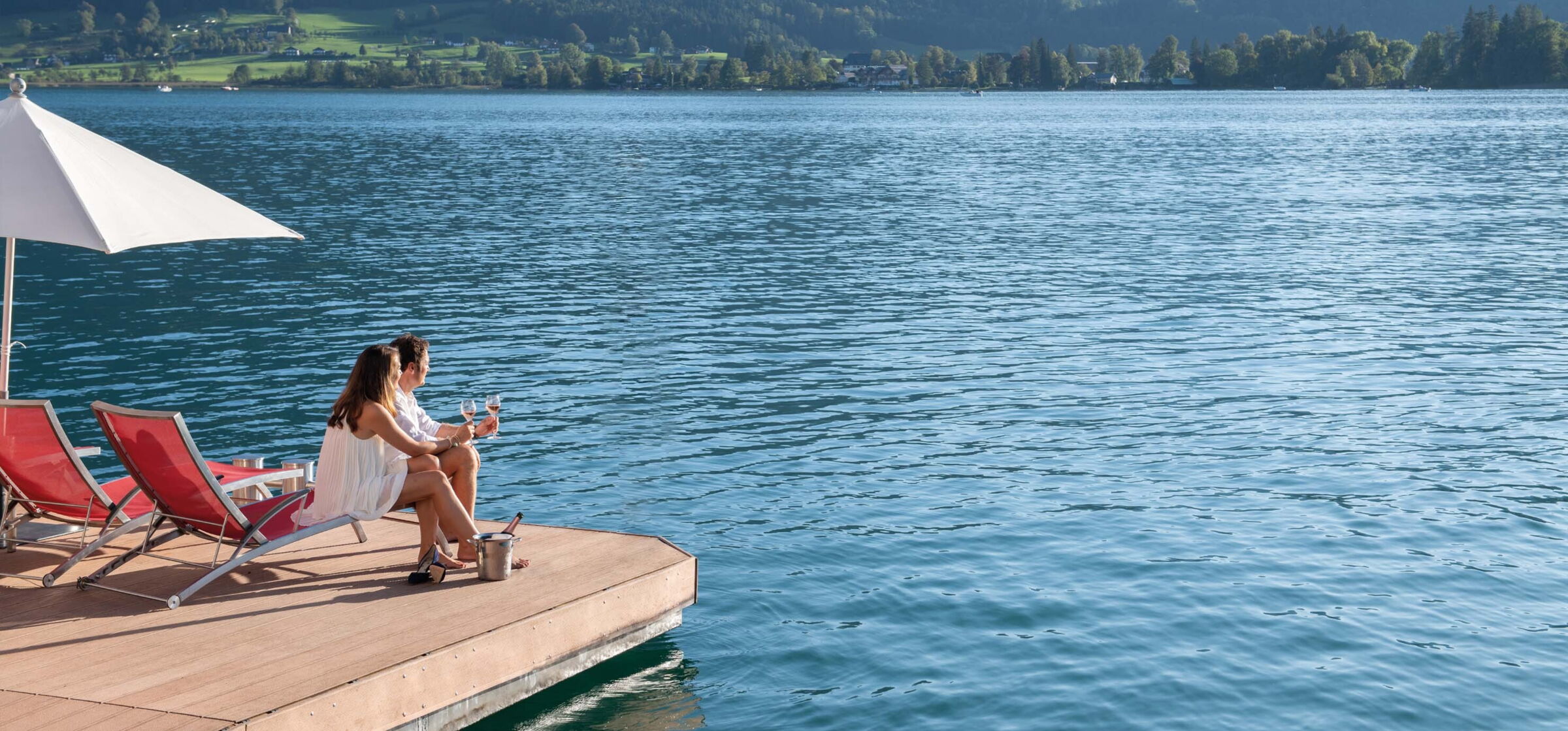 Couple holding wine glasses and sitting on loungers beside Lake Wolfgang