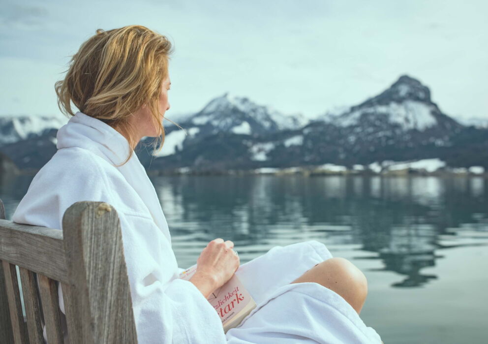 Eine Frau sitzt am Wolfgangsee im Bademantel und hält ein Buch