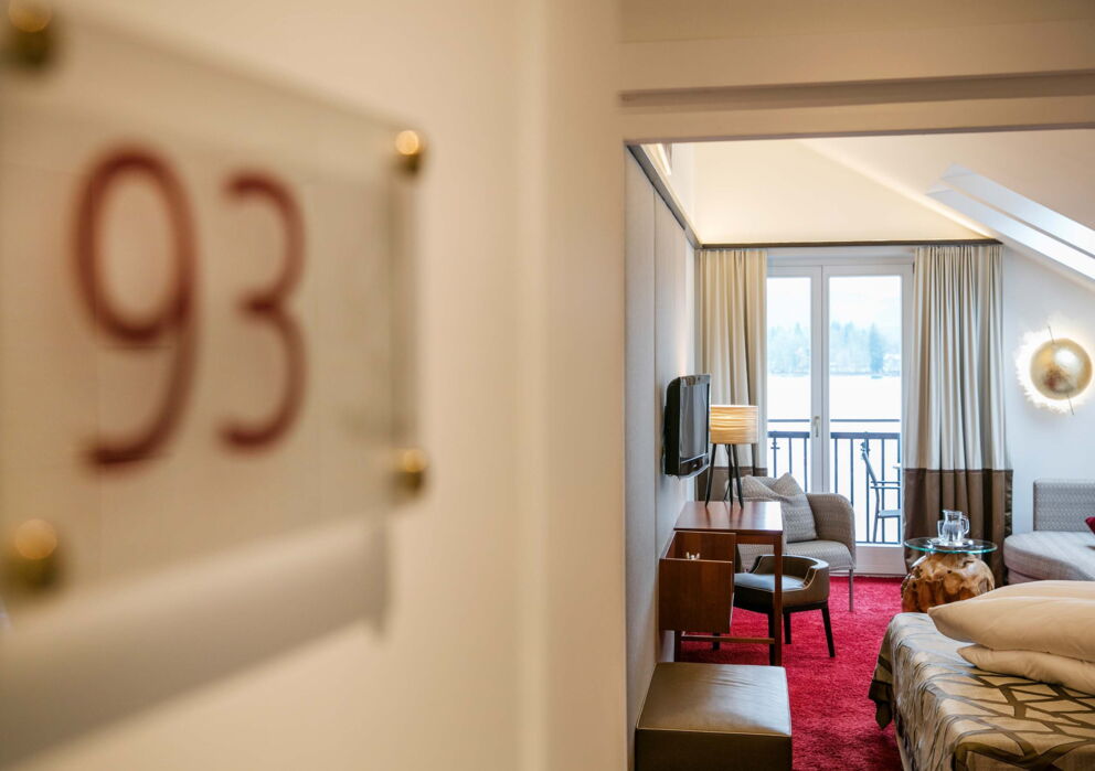 Bedroom with red carpeting as well as a balcony with a beautiful view of Lake Wolfgang, equipped with a TV, a double bed as well as a small living area with sofa, living room chair and table.