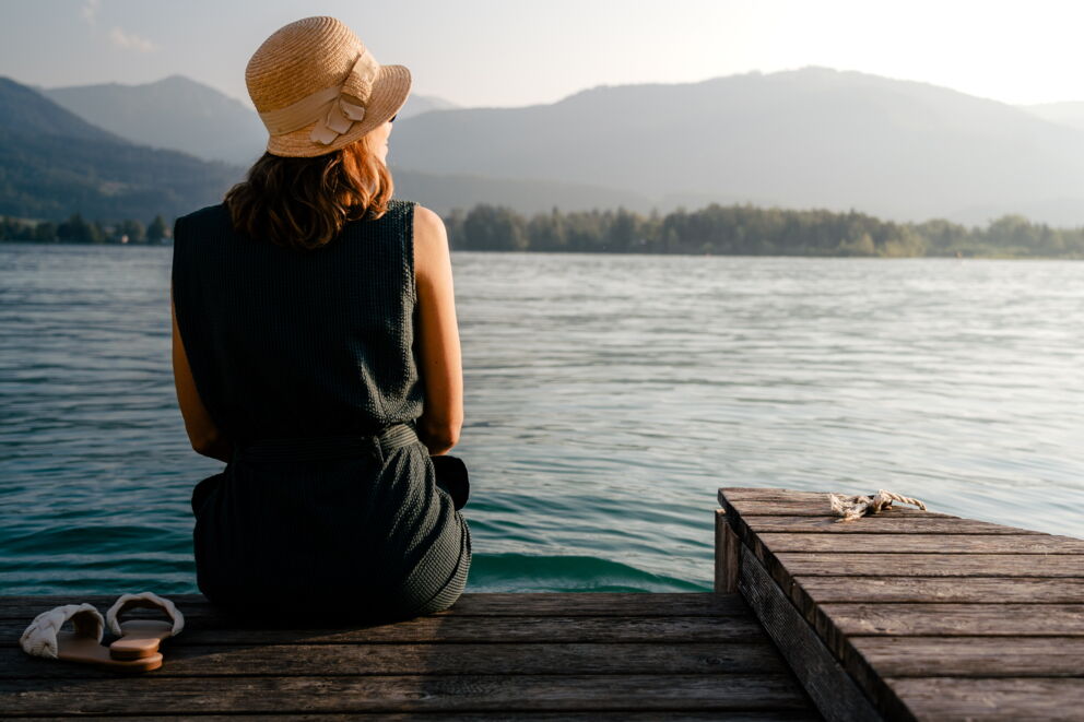Frau sitzt auf einem Holzsteg am See, mit Bergen im Hintergrund.