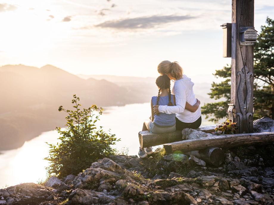 Eine mutter und ihre Tochter sitzen dem einem Gipfelkreuz und schauen auf den Wolfgangsee