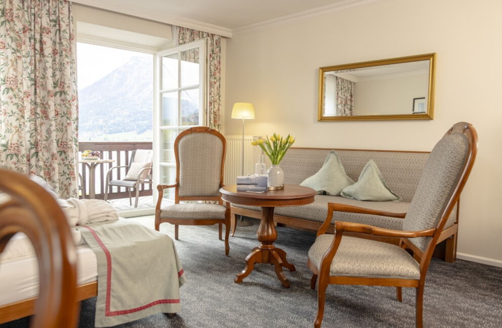 Sitzbereich im Romantik Hotel Im Weissen Rössl am Wolfgangsee mit Sofa, zwei Sesseln, rundem Tisch, Spiegel, floralen Vorhängen und Balkonzugang mit Blick auf den Wolfgangsee und die Berglandschaft.