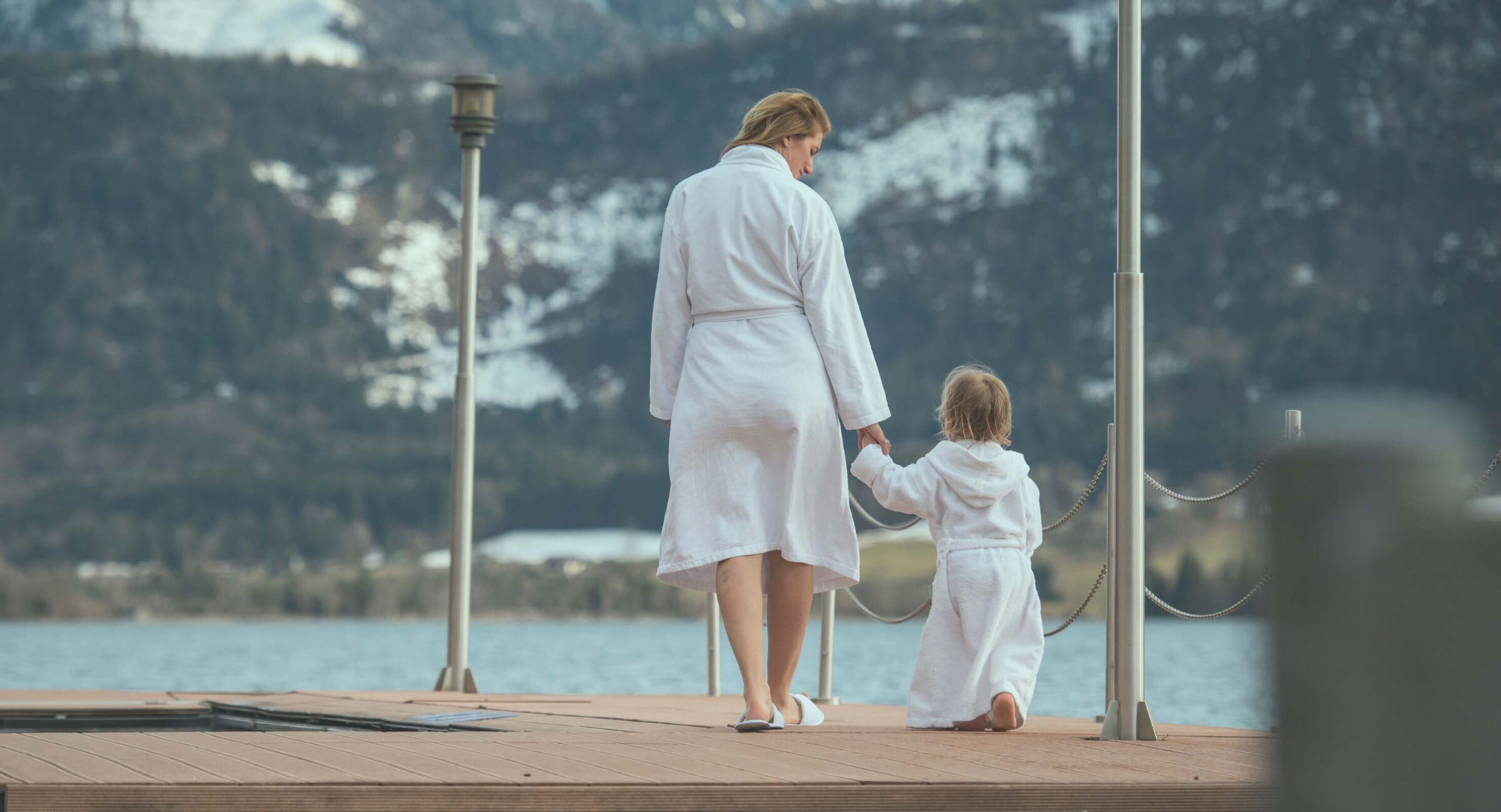 Eine Frau hält ihr kind an der Hand und geht über einen Holzsteg zum Wasser.