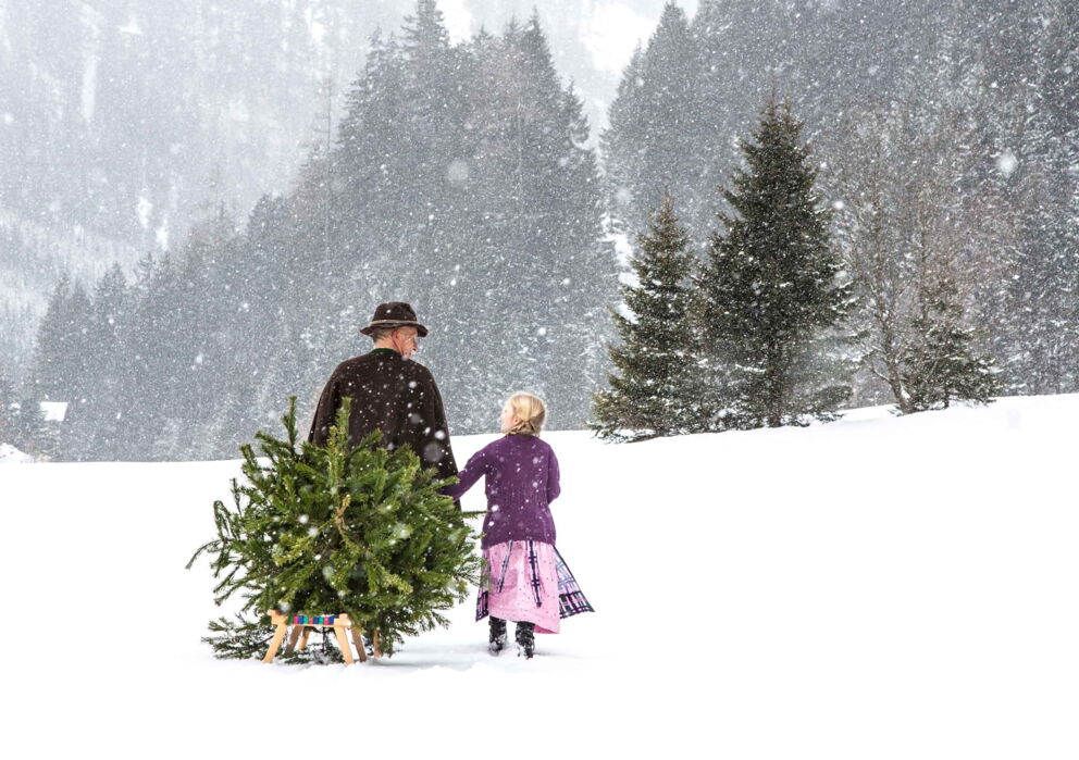Ein Mann und eine Mädchen gehen durch den Schnee, der Mann zieht einen Christbaum auf einem Schlitten