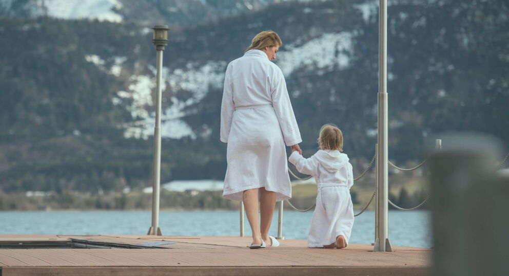 A woman holds her child by the hand and walks over a wooden deck to the water.
