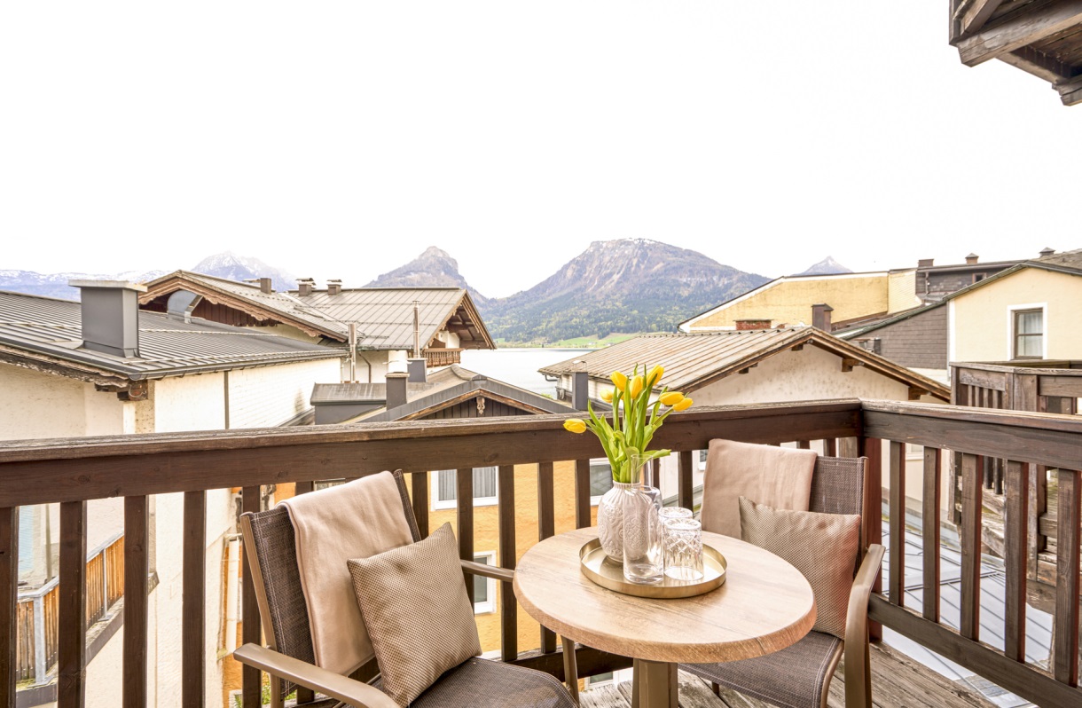 A balcony at the Romantik Hotel Im Weissen Rössl on Lake Wolfgang, featuring two armchairs, a small table, a wooden railing, and a view over the rooftops to Lake Wolfgang and the surrounding mountains.