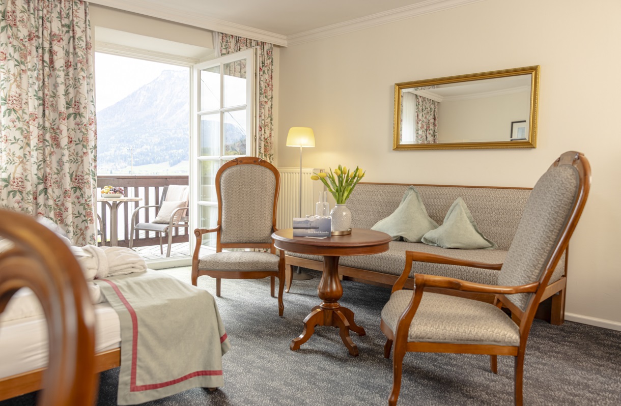 Sitzbereich im Romantik Hotel Im Weissen Rössl am Wolfgangsee mit Sofa, zwei Sesseln, rundem Tisch, Spiegel, floralen Vorhängen und Balkonzugang mit Blick auf den Wolfgangsee und die Berglandschaft.