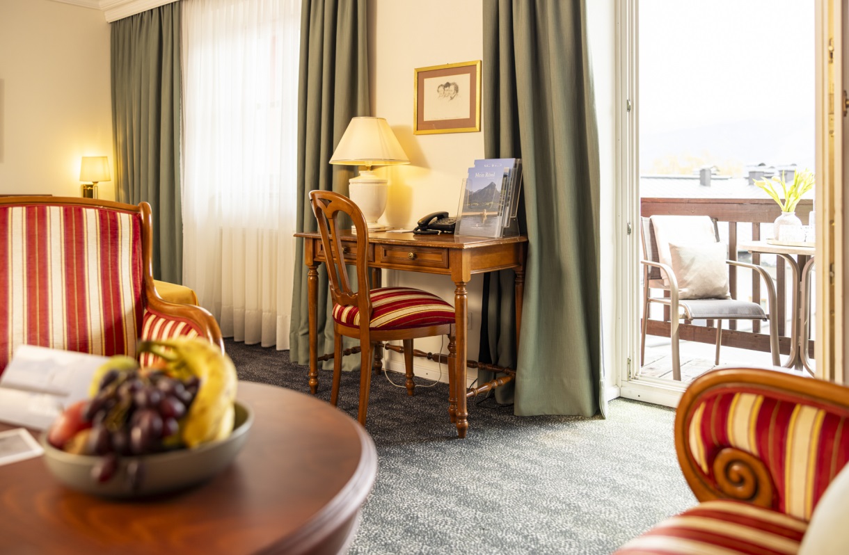 Sitzbereich im Romantik Hotel Im Weissen Rössl am Wolfgangsee mit Sesseln, rundem Tisch, Schreibtisch, Stuhl, Obstschale und Balkonzugang mit Blick auf den Wolfgangsee.