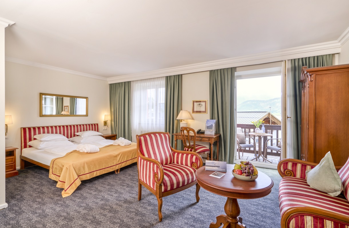 Doppelzimmer im Romantik Hotel Im Weissen Rössl am Wolfgangsee mit Doppelbett, Sitzbereich, Sesseln, rundem Tisch, Schreibtisch, Kleiderschrank und Balkonzugang mit Blick auf den Wolfgangsee.