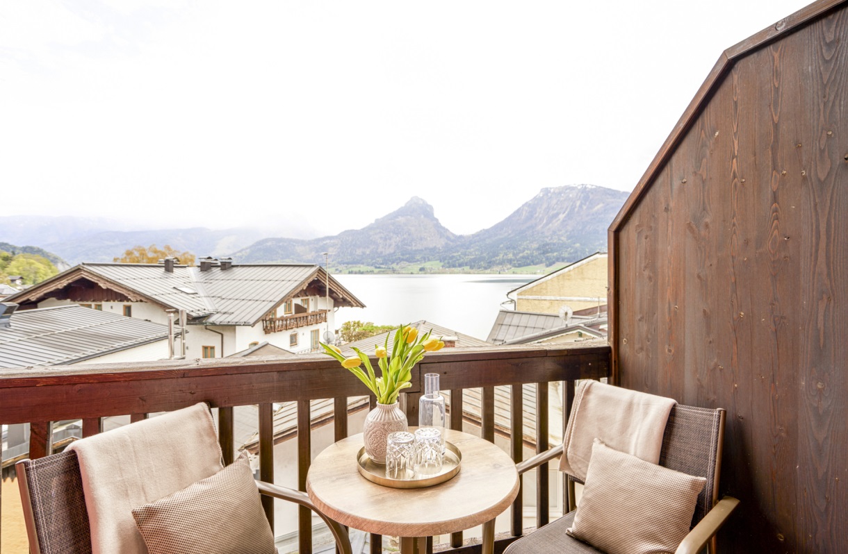 Balkon im Romantik Hotel Im Weissen Rössl am Wolfgangsee mit zwei Sesseln, kleinem Tisch, Holzgeländer und Blick über die Dächer zum Wolfgangsee sowie zur Berglandschaft.