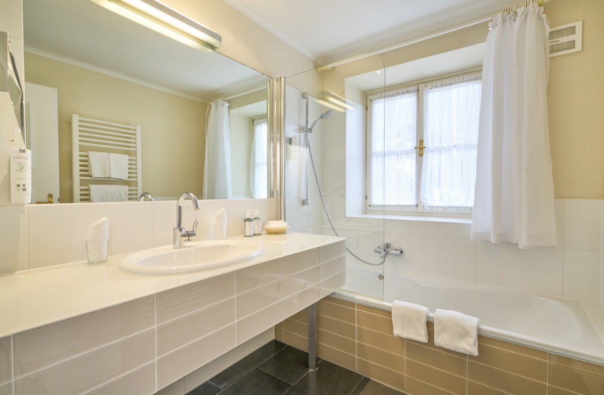 Badezimmer im Romantik Hotel Im Weissen Rössl am Wolfgangsee mit Badewanne, Duschvorrichtung, großem Waschtisch, Spiegel, Handtuchheizkörper und heller Fliesenausstattung.