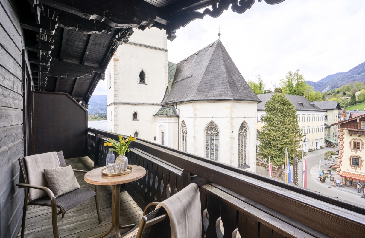 Balkon im Romantik Hotel Im Weissen Rössl am Wolfgangsee mit zwei Sesseln, kleinem Tisch, Holzgeländer und Blick auf die Kirche von St. Wolfgang sowie die umliegende Berglandschaft.
