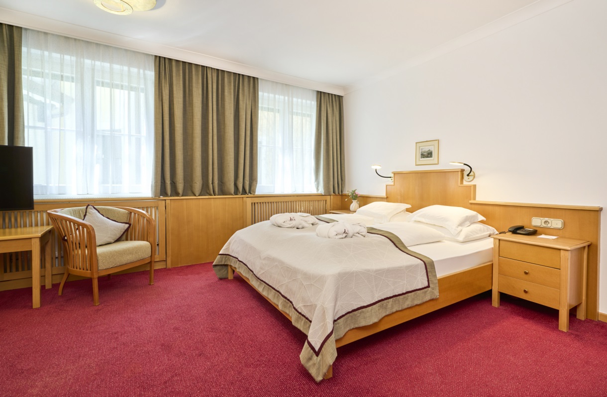 Familienzimmer im Romantik Hotel Im Weissen Rössl am Wolfgangsee mit Doppelbett, Fernseher, Sitzgelegenheit, Nachttisch, großen Fenstern, rotem Teppichboden und klassischer Holzausstattung.