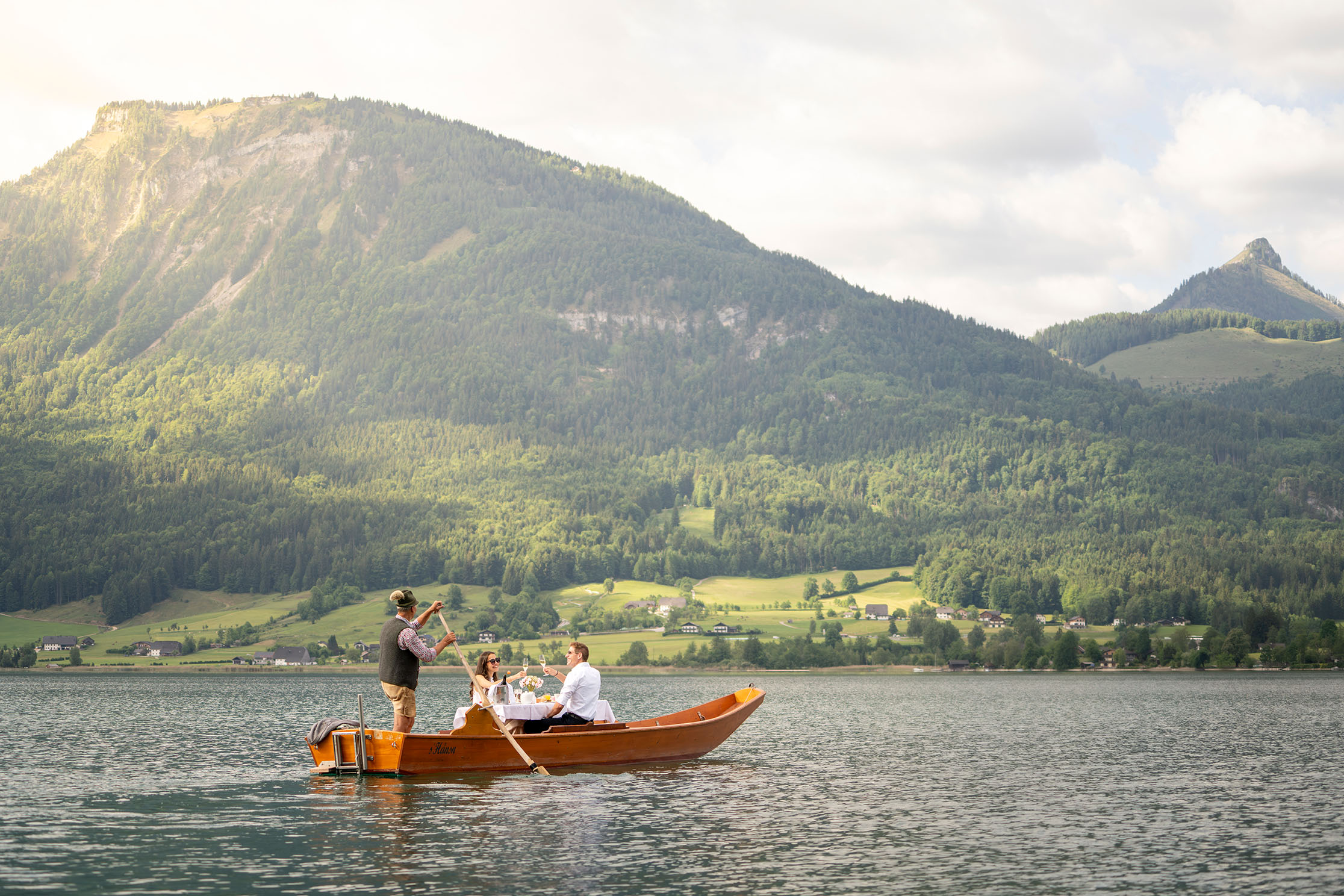 Two people in a wooden boat on Lake Wolfgang with green mountains in the background.